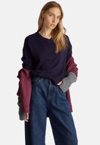 Suéter azul marino con escote en V, superpuesto con una sudadera color marrón, y puños acanalados en gris, acompañado de pantalones vaqueros de pierna amplia.