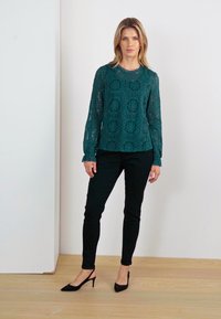 Teal csipke hosszú ujjú blúz virágmintával, fekete testhez álló nadrággal és fekete hegyes orrú magassarkú cipővel párosítva; sima textúrák és szabott fazon.