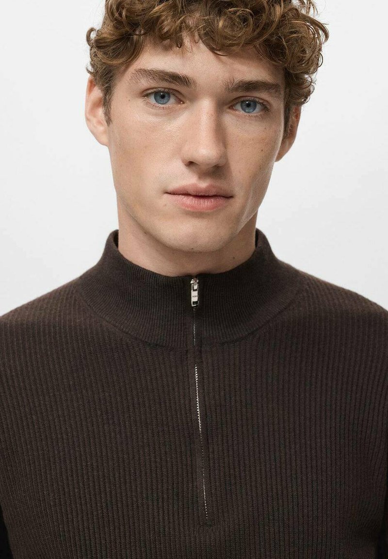 Brauner, gerippter Pullover mit hohem Kragen und einem Viertel-Reißverschluss. Die Stoffstruktur wirkt weich, mit einem glatten Reißverschlussdetail am Hals.