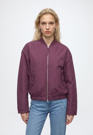 Blonde Frau mit welligem Haar, die eine lila Bomberjacke mit Reißverschluss und hellblaue Jeans trägt, steht vor einem schlichten hellen Hintergrund.