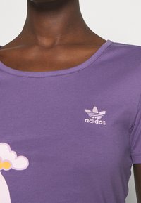 Fioletowa bawełniana koszulka z prążkowaną teksturą, dekoltem w serek oraz małym, białym, haftowanym logo Adidas na piersi. Posiada kolorowe szczegóły graficzne.