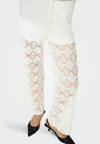 Pantaloni palazzo in pizzo bianco con un intricato motivo a diamante, dalla vestibilità ampia e con tessuto trasparente, abbinati a scarpe nere a punta.