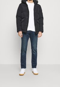 Giacca a vento nera con cappuccio, chiusura con zip e tasche laterali, indossata sopra un maglione bianco. Abbinata a jeans blu scuro e scarpe da ginnastica bianche.