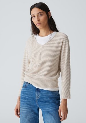 Beige gerippter Strickpullover mit V-Ausschnitt, langen Ärmeln, über einem weißen Hemd getragen, kombiniert mit blauen Jeans, die ein strukturiertes Design aufweisen.