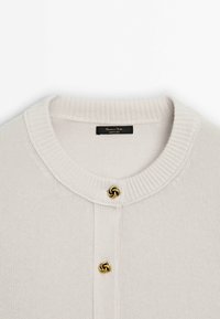 Valkoinen neulecardigan, jossa on pyöreä pääntie, ribbineuleiset hihansuut ja kaksi koristeellista kultasävyistä solmupainiketta edessä.