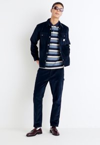 Giacca in velluto a coste blu navy sopra una polo a righe nei colori blu, crema e grigio. Pantaloni blu navy abbinati e scarpe eleganti bordeaux. Sfondo neutro.