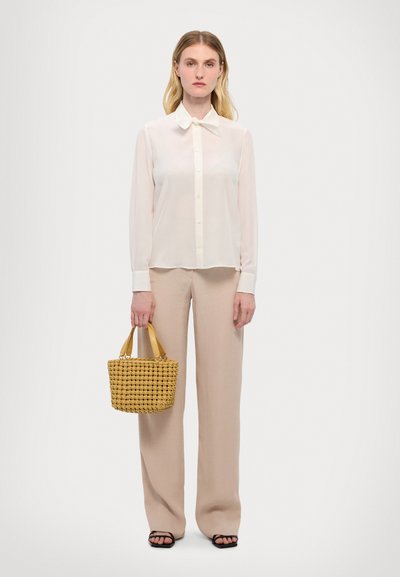 Transparente weiße Bluse mit langen Ärmeln, kombiniert mit hoch taillierten beigen Hosen. Trägt eine kleine geflochtene gelbe Handtasche. Schwarze Sandalen sind sichtbar.