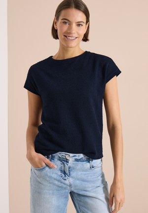 Femme aux cheveux bruns courts sourit, portant un haut à manches courtes bleu marine texturé et un jean bleu clair, une main dans la poche, fond neutre.