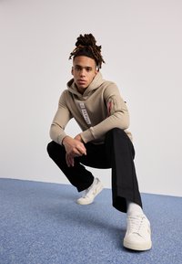 Beige hettegenser med glidelåsdetalj på siden og logo, sammen med sorte bukser og hvite sneakers. Bakgrunnen er lys grå med blått gulv.