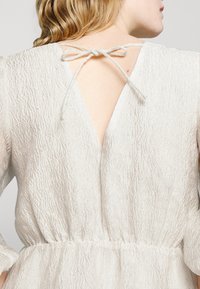 Robe beige clair texturée avec un dos en V et fermeture à nouer. Comprend des manches longues et une taille élastique pour une coupe décontractée.