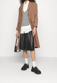 Manteau en laine marron porté sur une chemise blanche, gilet à motif pied-de-poule et jupe noire plissée. Chaussures à plateforme noires et petit sac à main.