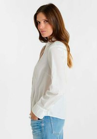 Wit shirt met lange mouwen, knoopsluiting bij de kraag, gladde textuur en één borstzak. Gecombineerd met lichtblauwe spijkerbroek, zijaanzicht.