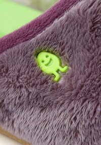 Zapatilla de casa morada y peluda con una superficie texturizada y un detalle de cara sonriente en un brillante color verde. El diseño es suave y acogedor, adecuado para uso en interiores.