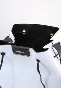 Biała torebka skórzana o fakturze z czarnym wnętrzem, czarnym sznurkiem ściągającym i metalową tabliczką z logo "GUESS" na zewnątrz.