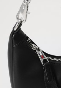 Sac en simili cuir noir avec fermeture éclair, quincaillerie argentée et une étiquette affichant le logo de la marque. Sangle d'accent avec une attache à clip.