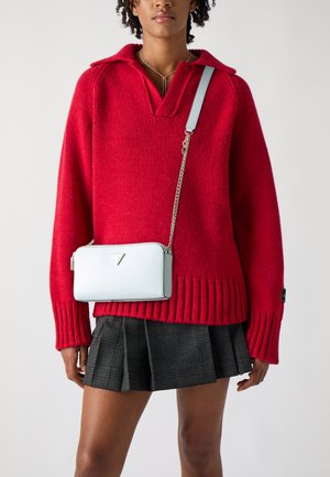 Suéter de punto rojo con escote en V y puños acanalados, combinado con un bolso bandolera azul claro que presenta un emblema en forma de triángulo y correa de cadena.
