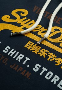 Marineblaues Sweatshirt mit gelbem und weißem, abblätterndem Text, ausgestattet mit cremefarbenen Kordeln und metallischen Spitzen. Der Text enthält Marken- und Designdetails.