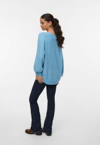 Pull bleu en maille avec une coupe ample, manches ballon et un décolleté en V. Associé à un jean évasé foncé et des bottines marron à talons.