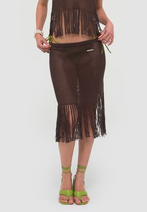 Kobieta nosząca brązową, dzianą spódnicę z frędzlami na zielonych majtkach bikini i zielonych sandałach na wysokim obcasie z paseczkami, trzymająca pas spódnicy.