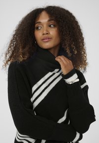 Moteris su garbanotais plaukais, vilkinti juodai-baltą juostuotą golfą, liečia apykaklę ir žiūri šone.