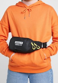 Persona que lleva una sudadera naranja y jeans azules, con una riñonera negra de Adidas que tiene tiradores amarillos en la cremallera y el logo en la parte frontal.