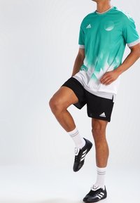 Turkos och vit gradient sporttröja med Adidas-logotyp, i kombination med svarta shorts. Modellen har på sig vita strumpor och svarta sportskor.