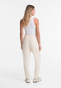 Light beige joggers with an elastic waistband and cuffed ankles, paired with a white tank top and beige sneakers. Smooth fabric texture.