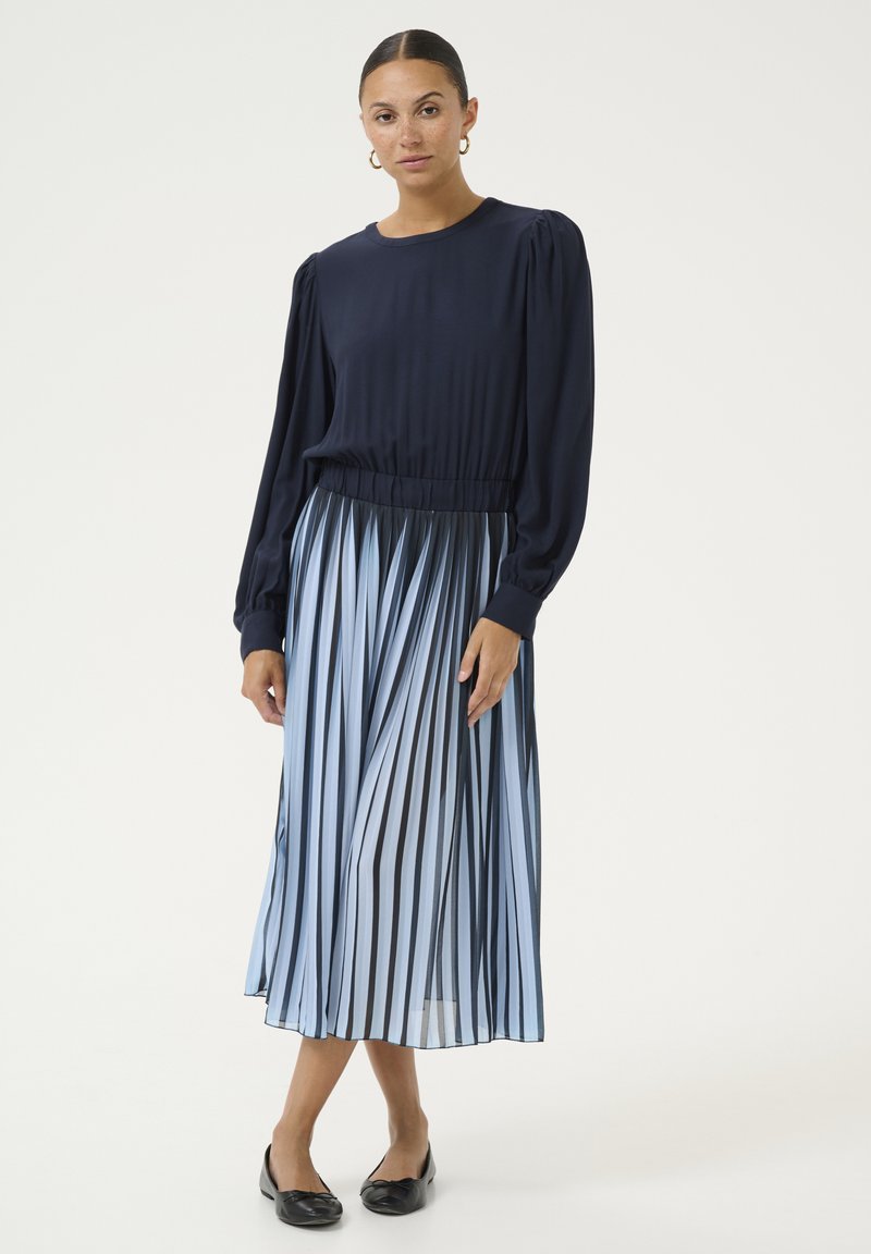 Tumedast sinine pluus puffvarrukatega, millele on sobitatud plisseeritud midi seelik, millel on vahelduvad sinised ja mustad triibud. Mustad lamedad kingad.