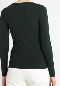 Ciemnozielony sweter z długim rękawem z prążkowanym dekoltem i obrębem. Gładka tekstura, dopasowany krój i minimalna ilość szwów, noszony z białymi spodniami.