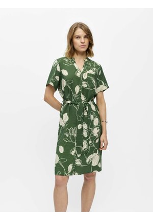 Femme portant une robe verte longueur genou avec des motifs floraux blancs et une ceinture en tissu, debout sur un fond clair uni.