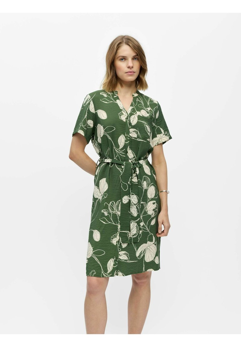 Femme portant une robe verte longueur genou avec des motifs floraux blancs et une ceinture en tissu, debout sur un fond clair uni.