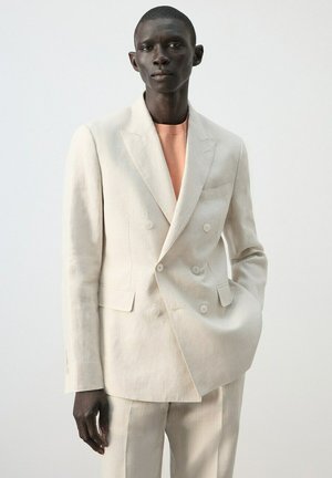 Uomo che indossa un abito doppiopetto crema su misura e una maglietta pesca con scollo tondo, in piedi con una mano in tasca davanti a uno sfondo semplice.