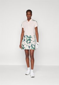 Ljusrosa pikétröja med korta ärmar och två knappar, kombinerad med en kjol i blommönster i grönt och vitt. Vit golfhandske och skor.