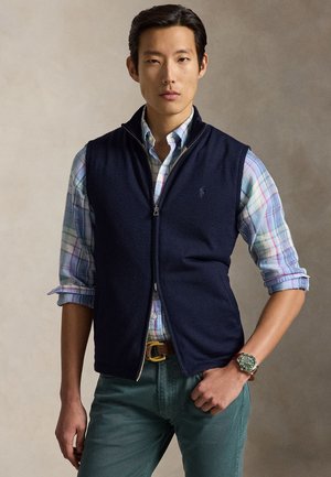 Muž nosiaci námornícky modrú vestu na zips cez vyhrnutú pastelovú kockovanú košeľu, zelené nohavice, hnedý opasok a náramkové hodinky na neutrálnom pozadí.