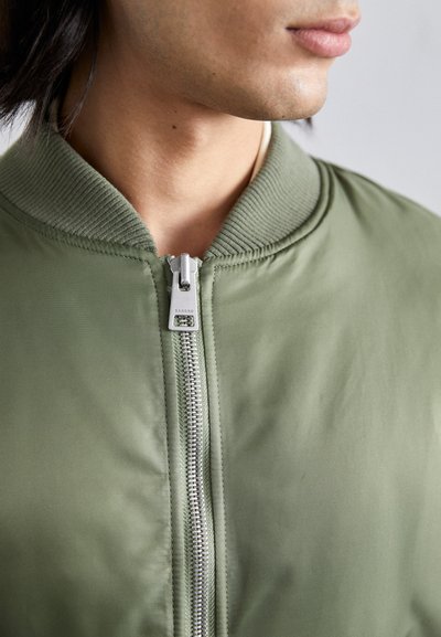 Chaqueta bomber verde oliva con cuello acanalado, con cremallera plateada y detalles de costura; textura de tela suave y ajuste ceñido.