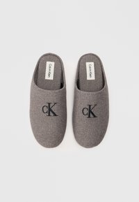 Papuci gri din fetru cu spate deschis, care prezintă un logo brodat negru 'CK'. Textură netedă cu căptușeală moale. Etichetele din interior citesc "Calvin Klein".