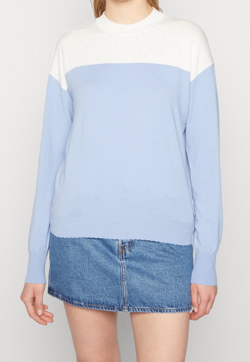 Femme portant un pull à manches longues en color-block blanc et bleu clair, associé à une mini-jupe en jean bleu, debout devant un fond uni.