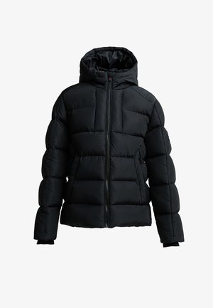 Teenager iført en sort hættejakke, mørkegrå joggingbukser, hvide sneakers, foran en ensfarvet lys grå baggrund.