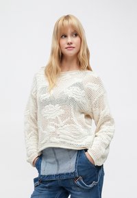 Pull en maille écru avec un design ample et ajouré, présentant des motifs floraux texturés. Associé à une salopette en denim, avec des détails de coutures visibles.