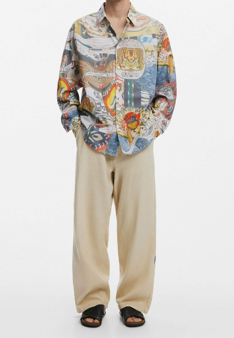 Personne portant un pantalon beige à jambes larges, une chemise à manches longues colorée avec des motifs abstraits, et des sandales noires ouvertes à l'avant.