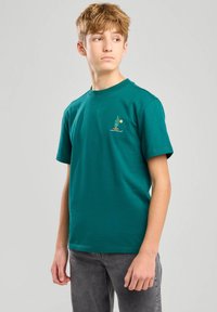 Camiseta de algodón verde con cuello redondo, mangas cortas y un pequeño gráfico de un cactus con un sol. La tela tiene una textura suave.