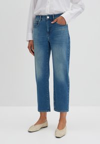 Jeans in denim azzurro chiaro con taglio dritto, cinque tasche e leggere scoloriture. Abbinati a slip-on beige e una camicia bianca con bottoni.