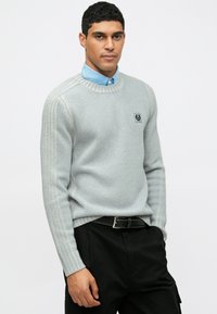 Ανοιχτό γκρι ριπτό πουλόβερ με στρογγυλή λαιμόκοψη, με μια μικρή ετικέτα λογότυπου. Φορεμένο πάνω από μπλε πουκάμισο με γιακά, συνδυασμένο με μαύρο παντελόνι.