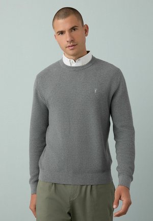 Suéter gris de punto con cuello redondo y puños acanalados, utilizado sobre una camiseta blanca con cuello. Presenta un sutil logo en el pecho.