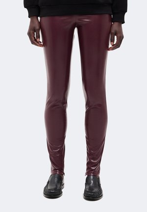 Personne portant un pantalon en cuir rouge foncé et des chaussures style mocassin noires, debout avec les mains détendues le long du corps.
