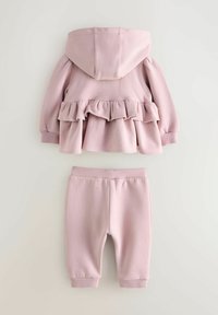 Mäkký ružový detský outfit s kapucňou, volánikovou bundou a zodpovedajúcimi nohavicami na gumu, položený na neutrálnom pozadí.