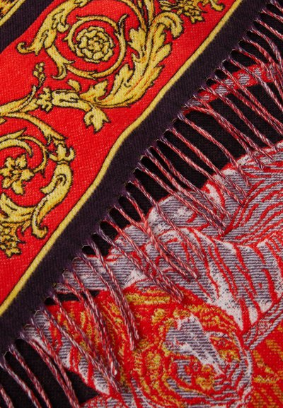 Close-up of a richly patterned fabric featuring gold floral scrolls on red, fringed edges, and intricate red, black, and gray woven details.