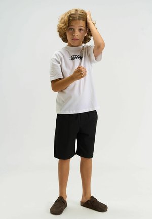 Biele tričko s krátkym rukávom s čiernym logom, v kombinácii s čiernymi šortkami a hnedými šmýkačkovými topánkami, na dieťati s kučeravými vlasmi.