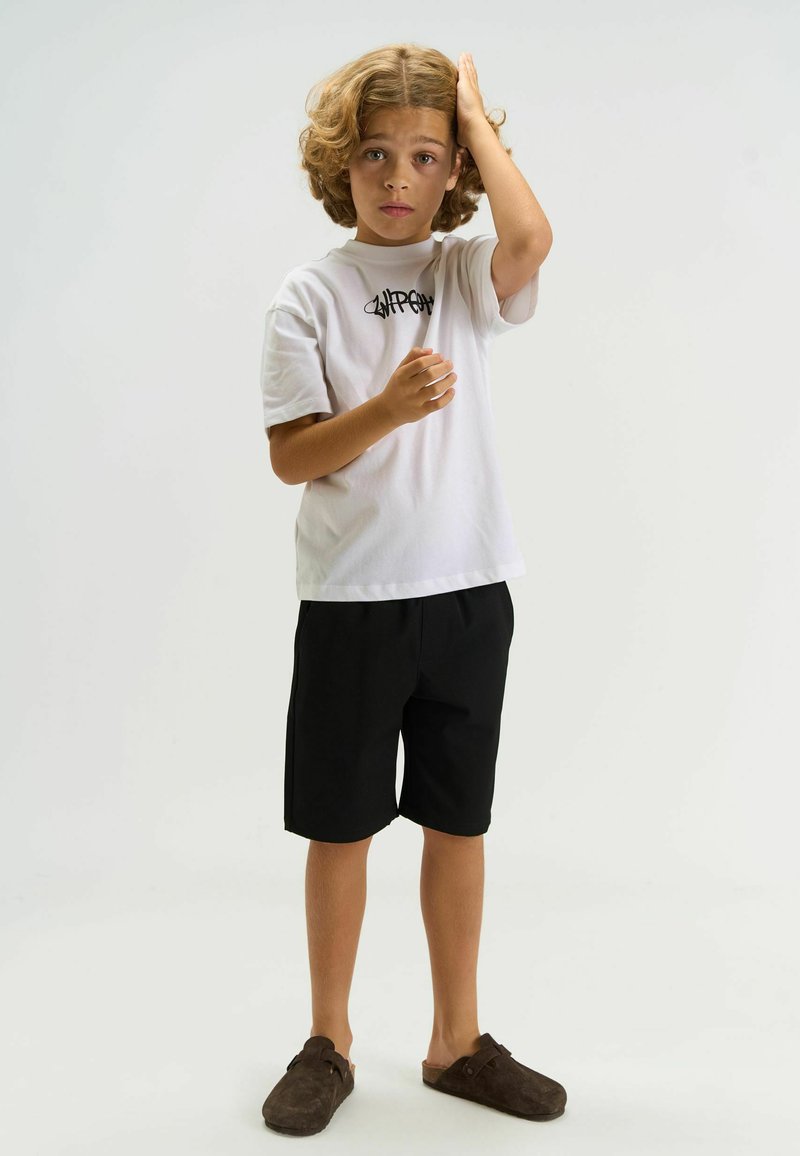 T-shirt blanc à manches courtes avec un logo noir, associé à un short noir et des chaussures marron à enfiler, porté par un enfant aux cheveux bouclés.