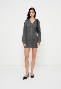 Γκρι πλεκτό φόρεμα με μακριά μανίκια, V-λαιμόκοψη και μπροστινά κουμπιά. Ζευγάρι μαύρες ψηλοτάκουνες γόβες. Μοντέλο κρατά τσάντα με μαύρο και λευκό πουά.